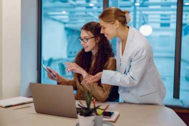 İki iş kadını, günümüz iş yerinin dinamiklerini sergilemek için modern bir ofiste dizüstü bilgisayarla iş birliği yapıyorlar.