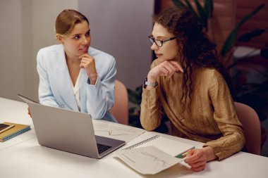 Modern bir çalışma alanında, iki kadın birlikte bir dizüstü bilgisayar üzerinde çalışırken canlı bir tartışmaya girişirler.