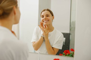 Neşeli bir kadın, parlak ve modern bir banyo ortamında kişisel cilt bakımını zevkle yapar.