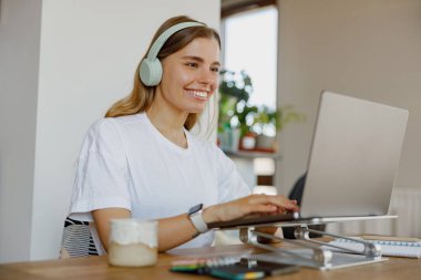 Neşeli genç bir kadın rahat ev ortamından bilgisayarını ve kulaklıklarını açık tutarak çalışmaktan zevk alıyor.