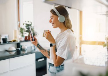 Genç bir kadın telefonunu kullanarak kahvesini içiyor ve neşeli, neşeli bir mutfakta müzik dinliyor.