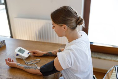 Bir kadın rahat ve sıcak iç mekanında dijital monitör kullanarak tansiyonunu dikkatlice ölçer.