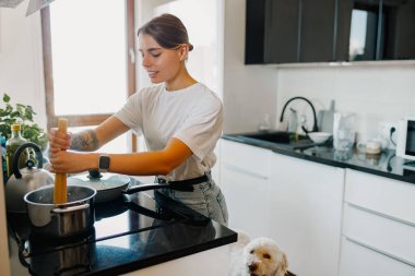 Önlüklü genç bir kadın, köpeği yakından izlerken çağdaş mutfağında demlik karıştırıyor.