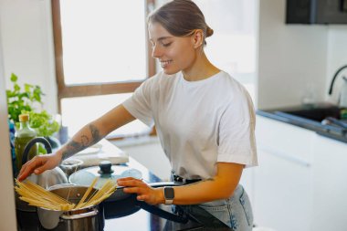 Genç bir kadın, parlak bir mutfakta makarna pişirmeyi, aşçılık becerilerini ve taze malzemelerini sergilemeyi sever..