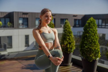 Bir kadın zarif bir şekilde, yemyeşil ve güzel şehir manzarasıyla çevrili bir çatıda yoga pozları veriyor.