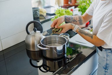 Bir insan, şık bir mutfaktaki kaynar suya taze otlar ekliyor. Mutfak yaratıcılığı ve tutku sergiliyor.