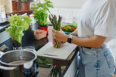 Bir insan taze otlarla dolu şık bir mutfakta sağlıklı yemekler hazırlarken kuşkonmaz tutar..