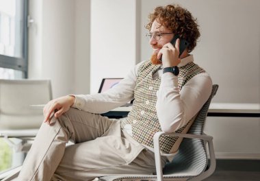 Kıvırcık saçlı genç bir adam şık, çağdaş bir ofis odasında otururken aktif olarak bir telefon görüşmesi yapıyor.