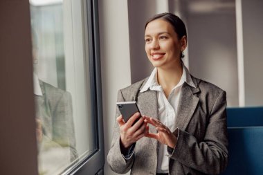 Profesyonel bir kadın akıllı telefonuyla meşgul, modern bir çalışma alanının penceresinde otururken parlak bir şekilde gülümsüyor.