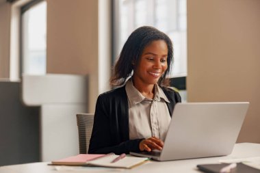 Profesyonel bir kadın gülümserken, parlak ve modern ofis ortamında dizüstü bilgisayarı üzerinde hevesle çalışıyor.