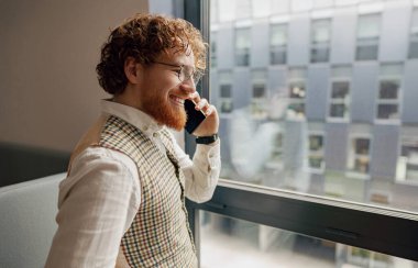 İyi giyimli bir adam, modern bir ofis ortamında telefonla konuşurken gülümsüyor.