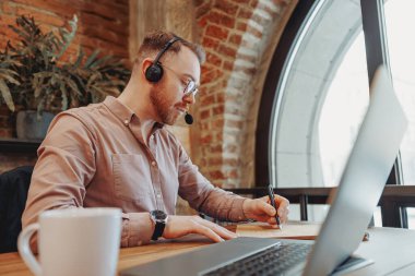 Odaklanmış bir adam şık bir kafede uzaktan kumandalı bir iş yapıyor, kulaklık takıyor ve yoğun bir şekilde not alıyor.