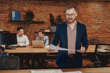 Bir adam, modern bir ofiste takım çalışması ve yaratıcılık sergileyen önemli belgelerle güvenle ayakta duruyor.