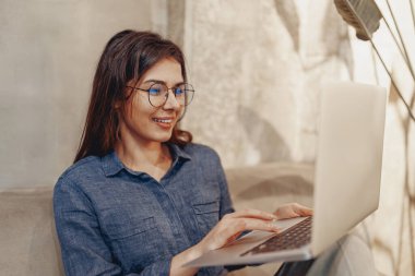 Diz üstü bilgisayarı üzerinde çalışırken mutlu bir şekilde gülümseyen bir kadın şık, rahat bir mekanda verimlilik için mükemmel.