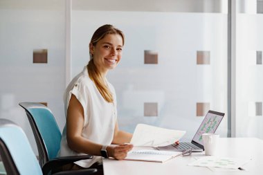 Neşeli ve profesyonel bir kadın modern bir ofis alanında çeşitli kağıtlar ve dizüstü bilgisayarla özenle çalışıyor.