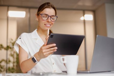 Parlak ve modern bir ofis çalışma alanında, genç bir kadın dikkatle büyük odaklanma ve konsantrasyonu olan bir tablet kullanıyor.