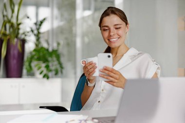 Parlak ve enerjik bir ofis ortamında neşeli bir kadın kahvesinin tadını çıkarırken akıllı telefonuna bakıyor.