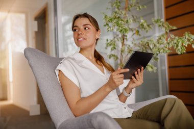 Genç bir kadın modern ofis ortamında bir tabletle meşgul olan şık bir sandalyede rahatça oturabilir.