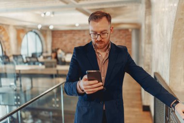 Modern bir çalışma alanında yer alırken akıllı telefonuyla meşgul olan şık ve iyi giyimli bir adam.