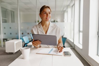 Kendini işine adamış ve işine odaklanmış profesyonel bir kadın hayat dolu ve parlak bir ofis ortamında tablet kullanıyor.