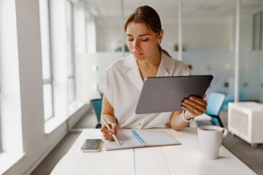 İşine dalmış profesyonel bir kadın. Parlak, modern ofis ortamında bir tablet ve not defteri kullanıyor.