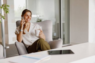 Moda kıyafeti giymiş şık bir kadın modern, şık bir ofis içinde telefonuyla neşe içinde sohbet ediyor.