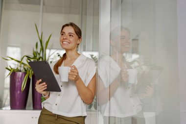 Neşeli bir kadın şık ve modern bir ofis ortamında tablet kullanırken keyifli kahvesinin tadını çıkarıyor.