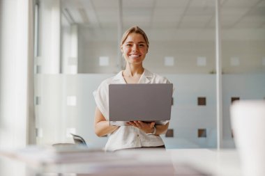 Neşeli genç bir kadın parlak ve modern ofis ortamında parlak bir dizüstü bilgisayar sunuyor.