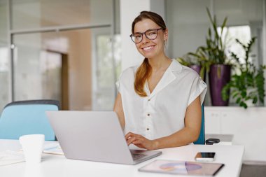 Gözlük takan kendine güvenen bir kadın, parlak ve modern bir ofis ortamında dizüstü bilgisayarıyla derinden meşguldür.