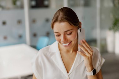 Neşeli ve gülümseyen bir kadın şık ve modern bir ofis ortamında telefon görüşmesi yapar.