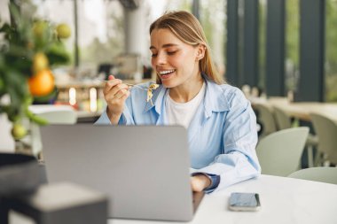 Neşeli bir kadın yemek yiyerek ve laptopuyla modern bir kafede çalışarak birden fazla iş yapar.