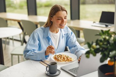 Modern ve modaya uygun bir kafe ortamında, genç bir kadın laptopuyla çalışırken sağlıklı bir kahvaltı yapıyor.