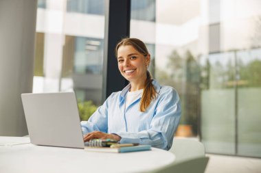 İyi aydınlatılmış, modern ve şık bir ofis ortamında özenle dizüstü bilgisayarı üzerinde çalışan neşeli bir kadın.