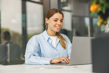 Gülümseyen bir kadın dizüstü bilgisayarıyla, uzak çalışma kültürünü temsil eden şık ve modern bir ofis ortamında meşgul.