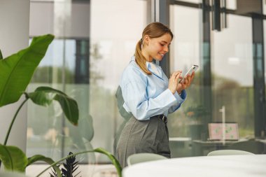 Modern, şık bir ofis ortamında otururken akıllı telefonuyla meşgul olan genç bir kadın.