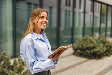 Şık mavi bir gömlek giyen gülümseyen genç bir kadın modern camdan bir binanın önünde elinde bir defter tutuyor.
