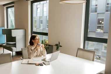 Dizüstü bilgisayarı üzerinde çalışırken derin düşüncelere dalmış bir kadının yer aldığı çağdaş bir ofis sahnesi.