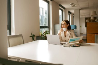 Bir kadın kahvesini yudumlarken titizlikle önemli belgeleri gözden geçirir. Parlak, çağdaş bir çalışma alanında.