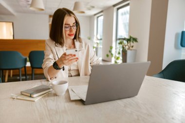 Kendini adamış bir kadın şık bir kafe atmosferinde otururken bilgisayarında ve akıllı telefonunda etkili bir şekilde birden fazla iş yapıyor.