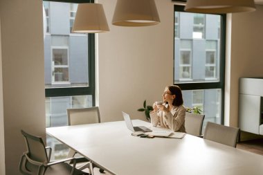 Bir kadın, çağdaş bir ofis ortamında otururken sakince ve düşünceli bir şekilde lezzetli bir içeceğin tadını çıkarıyor.