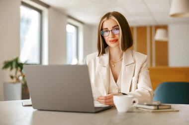 Odaklanmış genç bir kadın şık ofisinde dizüstü bilgisayarında, yanında kahve fincanıyla ustaca yazıyor.