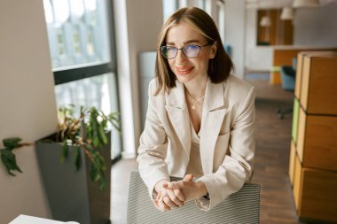 Gülümseyen bir iş kadını, modern bir ofiste kendine güvenerek duruyor, profesyonelliğini ve duruşunu sergiliyor.