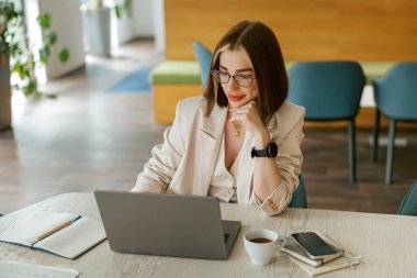 Şık bir spor ceket giymiş odaklanmış bir kadın, yaratıcı şeylerle dolu, parlak ve modern bir ofiste dizüstü bilgisayarıyla meşgul oluyor.