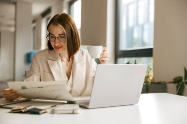 Bir kadın, yaratıcılık için tasarlanmış parlak ve modern çalışma alanında kahvesinin tadını çıkarırken bir kitaba çok dalmıştır.