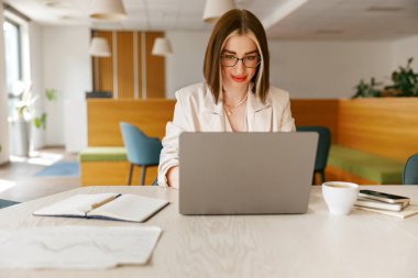 Kendini işine adamış ve odaklanmış genç bir kadın, şık bir ofis ortamında dizüstü bilgisayarı üzerinde çalışıyor.