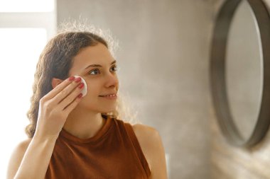 Genç bir kadın şık bir banyo ortamında neşeli yansımasına gülümserken mutlu bir şekilde deri altlığı kullanıyor.