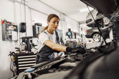 Kendini işine adamış ve odaklanmış bir kadın tamirci modern bir oto tamirhanesinde araba motorunu özenle inceliyor.