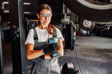 Yetenekli ve kendine güvenen bir kadın tamirci, çağdaş bir garaj ortamında elinde elektrikli bir aletle gururla durur.