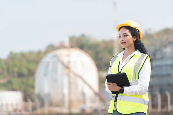 female engineer with hardhat with petrochemical factory background. asian woman holding tablet, plan and Walkie Talkie.
