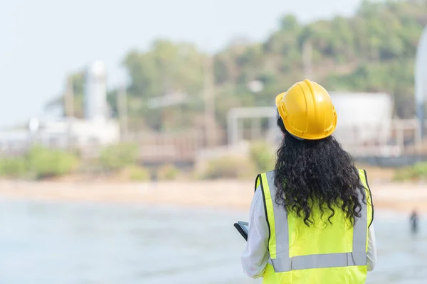 female engineer with hardhat with petrochemical factory background. asian woman holding tablet, plan and Walkie Talkie.